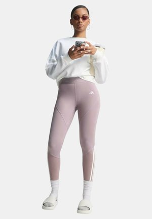 Persona con felpa bianca, leggings Adidas color malva, calzini bianchi, ciabatte, occhiali da sole rossi, che tiene uno smartphone, in piedi davanti a uno sfondo semplice.