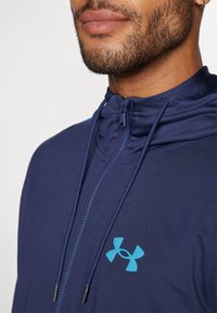 Marineblå zip-up jakke med høy krage, med et lyseblått logo på brystet og strukturert stoff for pusteevne.