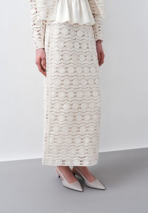 Vrouw draagt een witte midi rok van kant en een bijpassende kanten top met lange mouwen, gecombineerd met lichtgrijze slingback hakken met een puntige neus.