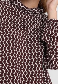 Burgundowa bluzka z białym wzorem w diamenty, miękka tkanina, długie rękawy i złożony kołnierzyk.