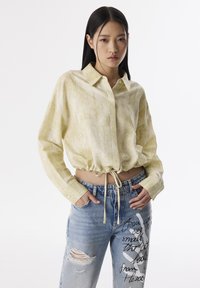 Lichtgele, gestructureerde, lange mouw shirt met kraag en striktaille. Gepaard met distressed blauwe spijkerbroek met bedrukte accenten.