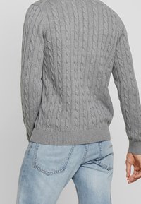 Szary sweter w splot warkoczowy z prążkowanymi mankietami i dolnym wykończeniem, noszony z jasnoniebieskimi dżinsami. Tekstura jest miękka, a design zawiera wypukłe przeszycia.