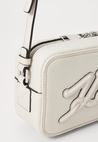 Borsa bianca in pelle con una finitura texturizzata, forma rettangolare, chiusura con zip e una tracolla corta. Presenta un distintivo design impressa sulla parte anteriore.