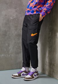 Černé cargo kalhoty s oranžovým logem Nike, doplněné bílými a hnědými teniskami s fialovými tkaničkami. Oblečení doplňuje svetr s barevným vzorem.