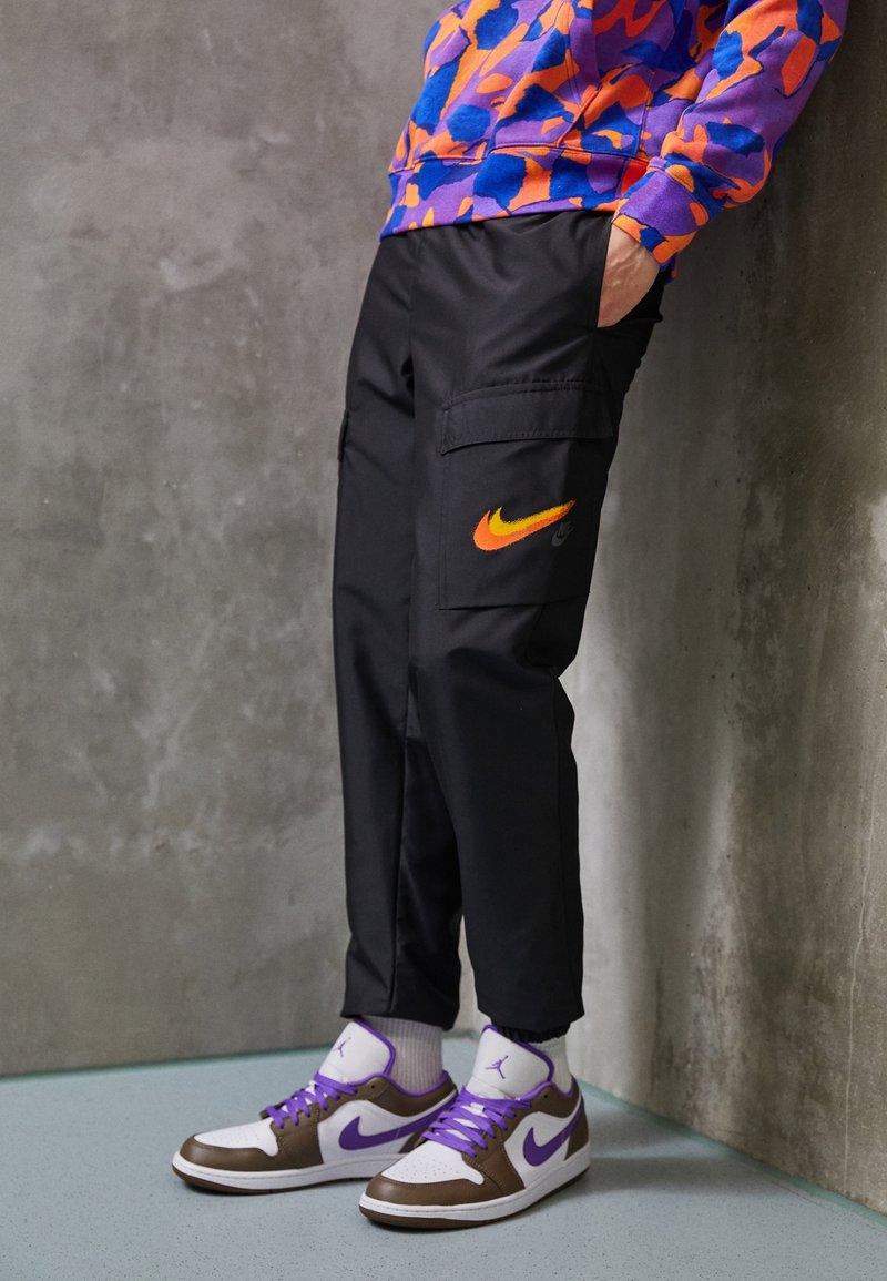 Černé cargo kalhoty s oranžovým logem Nike, doplněné bílými a hnědými teniskami s fialovými tkaničkami. Oblečení doplňuje svetr s barevným vzorem.