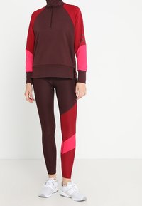 Sudadera de manga larga de medio cierre en color burdeos y rosa, con mangas raglán, combinada con leggings burdeos y rojos que presentan acentos geométricos en rosa.