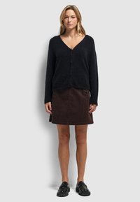 Zwarte gebreide cardigan met een V-hals en drie knopen, gecombineerd met een bruine ribfluwelen rok. Model draagt zwarte schoenen. Eenvoudig, gestructureerd ontwerp.