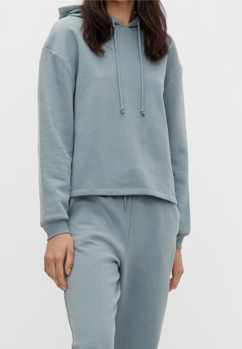Femme portant un sweat à capuche bleu clair avec cordons de serrage et un pantalon de survêtement assorti sur un fond uni.