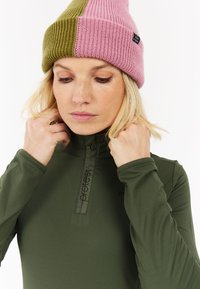 Groene langelijf basislaag met een ritskraag en logodetail. Het model draagt een kleurrijke gebreide muts in roze en groen.