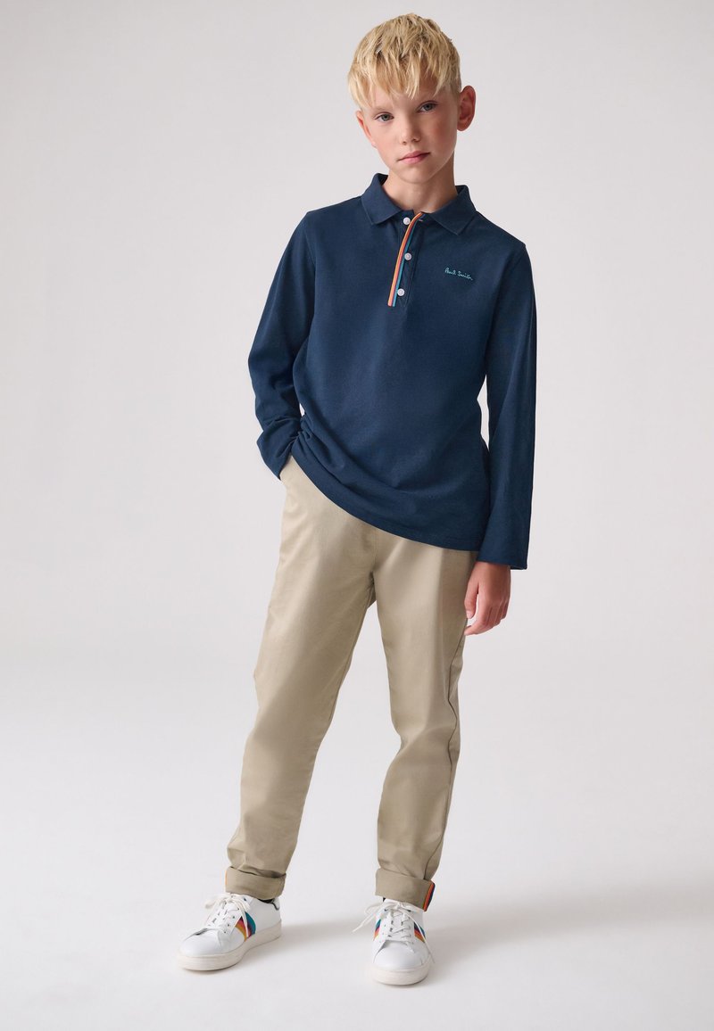 Tengerészkék hosszú ujjú galléros póló háromgombos gombolattal és narancssárga díszítéssel, bézs chino nadrággal és fehér, színes részletekkel ellátott tornacipővel párosítva.