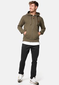Sweat à capuche zippé vert olive avec cordon de serrage, poches avant et patch logo. Associé à un pantalon noir et des chaussures de sport. Texture lisse.