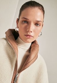 Giacca in pile con esterno crema e colletto in suede marrone, con cerniera frontale e dolcevita beige a coste sotto. Orecchini ad anello in argento.
