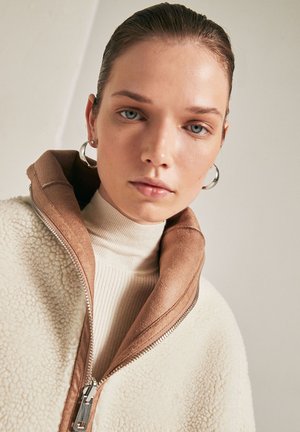 Giacca in pile con esterno crema e colletto in suede marrone, con cerniera frontale e dolcevita beige a coste sotto. Orecchini ad anello in argento.