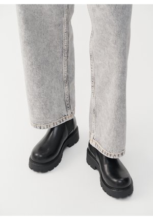 Personne portant un jean gris clair et des bottines noires à semelle épaisse, debout sur une surface blanche.