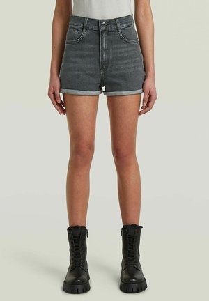 Donna che indossa shorts di jeans neri risvoltati, top bianco e stivali da combattimento neri con lacci, in piedi su uno sfondo semplice.