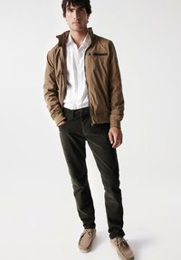 Chándal ligero marrón con cremallera sobre una camisa blanca, combinado con pantalones verde oscuro y zapatos beiges de cordones. Tejido suave, ajuste casual.