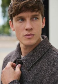 Brun struktureret jakke med krave, knaplukning og subtilt mønster. Modellen justerer jakken med den ene hånd.