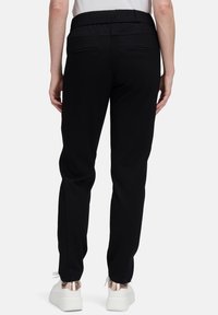 Osoba ubrana w czarne zwężane spodnie i białe trampki z metalicznymi różowymi złotymi akcentami, stojąca na jednolitym tle.