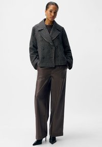 Doppelreihige Jacke mit einem strukturierten Schwarz- und Creme-Muster, ausgestattet mit einem breiten Kragen. Kombiniert mit hochgeschnittenen, glänzenden braunen Hosen und schwarzen Absätzen.