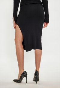 Schwarzer gerippter Strickrock mit hohem Schlitz, kombiniert mit schwarzen spitzen Absatzschuhen, die metallische Nieten an den Seiten haben.
