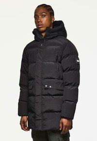 Schwarze Steppjacke mit quilteter Textur, Reißverschluss und Druckknopfverschlüssen, Kapuze und Seitentaschen. Mit einem Logo-Patch am Ärmel.