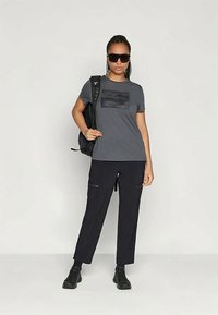 Mulher usando óculos de sol escuros, t-shirt gráfica cinza, calças pretas, botas pretas e carregando uma mochila preta, de pé contra um fundo liso.