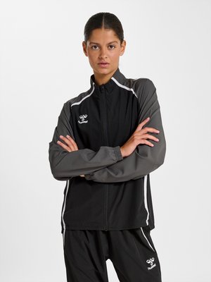 Frau mit dunklen Haaren in einem schwarzen und grauen Hummel Trainingsanzug, mit verschränkten Armen, nach vorne blickend vor einem schlichten weißen Hintergrund.