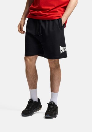 Mężczyzna ubrany w czarne szorty z logo na lewej nodze, białe skarpetki, czarne trampki i czerwoną koszulkę, stoi z jedną ręką w kieszeni.