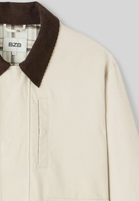 Veste extérieure beige avec un col en velours côtelé marron, dotée d'une poche poitrine et d'une doublure en tartan. Texture lisse, design structuré. Étiquette de la marque visible.