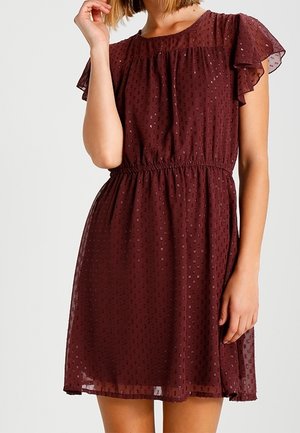 Kobieta ubrana w bordową sukienkę do kolan z krótkim rękawem, z taliowaniem i fakturowanym wzorem w grochy.