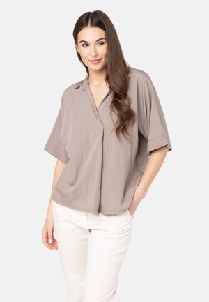 Blusa corta beige chiaro con colletto, vestibilità rilassata e un sottile dettaglio plissé sul davanti. Abbinata a pantaloni color crema.