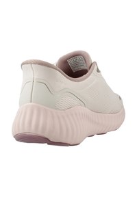 Zapatilla deportiva de color beige claro con parte superior de malla texturizada, suela redondeada en color rosa y sutiles detalles de acento. Diseño flexible y acolchado.