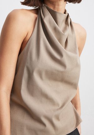 Donna che indossa un top beige senza maniche con una morbida scollatura drappeggiata su uno sfondo chiaro uniforme.