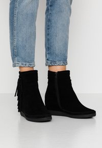 Černé zamšové kotníkové boty na klinu s třásněmi, bočním zipem a texturovanou podrážkou, v kombinaci s ohrnutými světle modrými džíny.