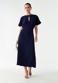 Sotilasnavy sininen, lattian pituinen mekko, jossa on lyhyet ja leveät hihat sekä avonainen kaula-aukko. Suora pinta, yhdistettynä beigeihin nauhapohjaisiin korkokenkiin ja valkoiseen käsilaukkuun.