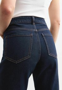 Jean en denim bleu foncé avec une taille haute ; comporte deux poches arrière avec des coutures orange et une taille visible. Texture lisse.