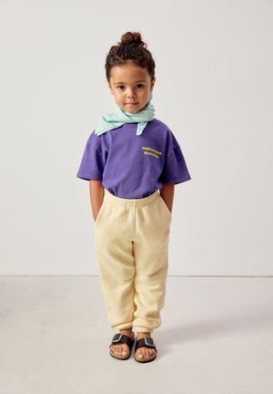Bambino con capelli ricci raccolti, indossa una maglietta viola "American Vintage", pantaloni della tuta beige, sandali neri e una sciarpa verde chiaro intorno al collo.