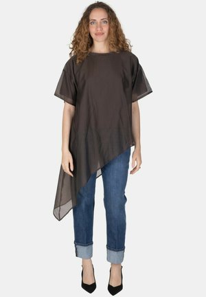 Donna con capelli ricci che indossa una maglia nera trasparente asimmetrica, jeans blu risvoltati e tacchi alti neri, in piedi su uno sfondo bianco.