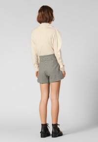 Vaaleanbeige pitkähihaisessa paidassa ja musta-valkoisissa houndstooth-shorteissa. Mallilla on mustat nilkkurit, joissa on koristeellinen hihna. Takakuvasta.