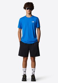 Blå kortärmad skjorta med en vit logotyp, svarta shorts, svarta skor och vita strumpor med logomönster. Lockigt hår och solglasögon.