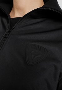 Zwart jack met ritssluiting en een gladde textuur, voorzien van een driehoekig zwart rubberen logo met de tekst "Rossignol" op de borst.