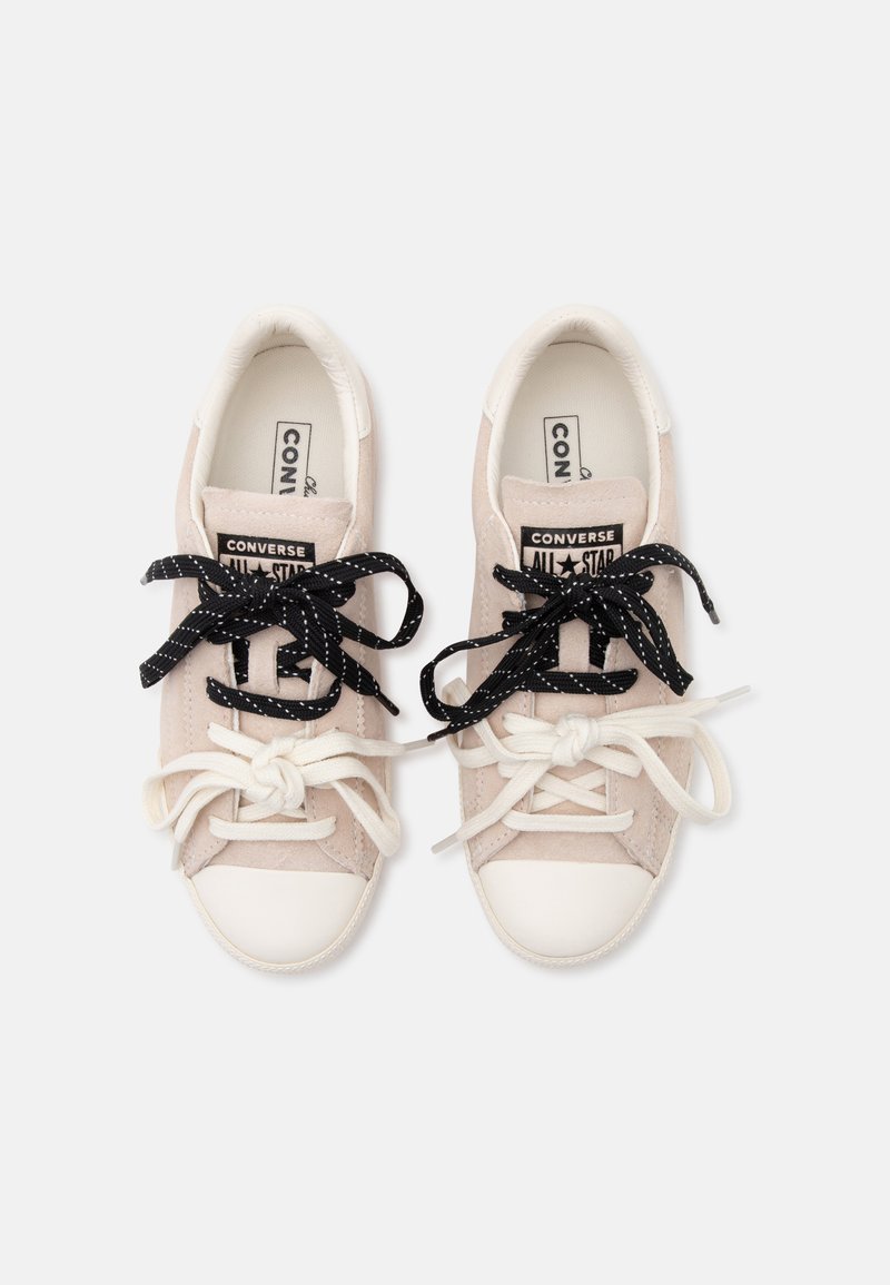 Paar beige Converse All Star Sneaker mit weißen Zehenkappen und verschiedenfarbigen schwarzen und weißen Schnürsenkeln, die zu Schleifen gebunden sind, auf weißem Hintergrund.