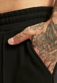 Black jogger pants with an elastic waistband. A hand is placed in the side pocket, showing detailed tattoos on the fingers and palm.