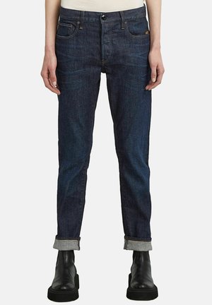 Jeans en denim bleu foncé avec une coupe fuselée, présentant des ourlets roulés, un style classique à cinq poches et une fermeture zippée.