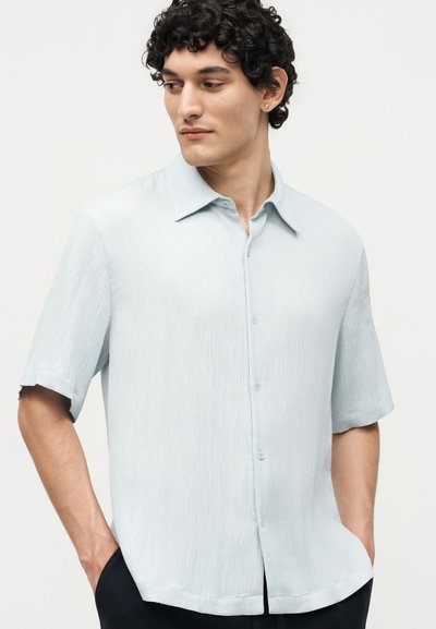 Νέος άνδρας με σγουρά μαλλιά, φορώντας ένα ανοιχτό μπλε κοντομάνικο πουκάμισο με υφή και κουμπιά, με τα χέρια στις τσέπες, κοιτάζοντας πλάγια.