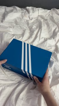 Kék kartondoboz fényes felülettel, három párhuzamos fehér csíkkal és „adidas” logóval az oldalán.