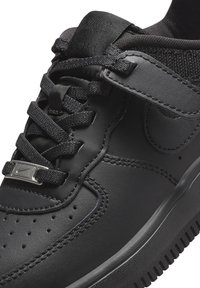 Gros plan d'une basket noire Nike Air Force 1 montrant une tige en cuir, des lacets texturés avec logo en métal et des détails cousus.