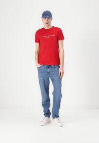 Camiseta de algodón roja con texto de logo blanco, combinada con jeans de mezclilla azules y zapatillas blancas. El modelo lleva una gorra azul clara, de pie contra un fondo liso.