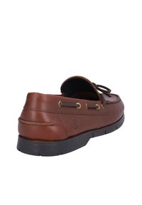 Zapato de estilo mocasín de cuero marrón con punta redondeada, superficie texturizada, suela oscura en contraste y cordones decorativos en los laterales.