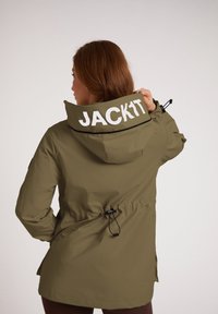 Oliwkowa wodoodporna kurtka z regulowanym kapturem, czarnymi akcentami, ściągaczem w talii oraz dużym białym napisem "JACK1T" na plecach.